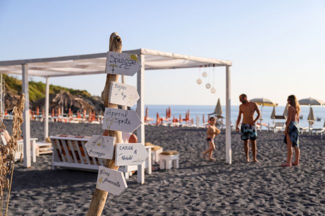 Spiaggia con sabbia scura, cartelli e persone in lontananza.