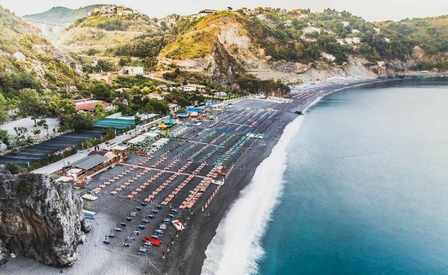 Spiaggia di ciottoli con file di ombrelloni, mare azzurro e colline verdi sullo sfondo.