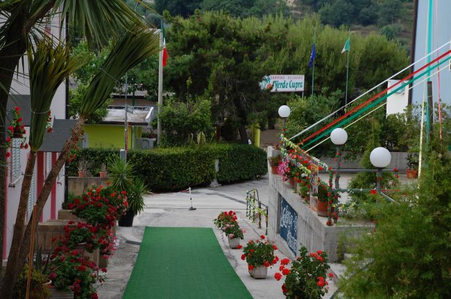 Un ingresso con passerella verde, piante fiorite ai lati e bandiere in lontananza.