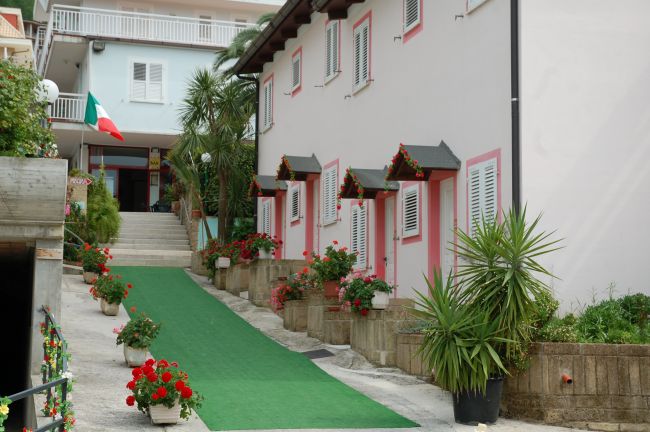Viale con piante e fiori, edificio rosa e bandiera italiana.