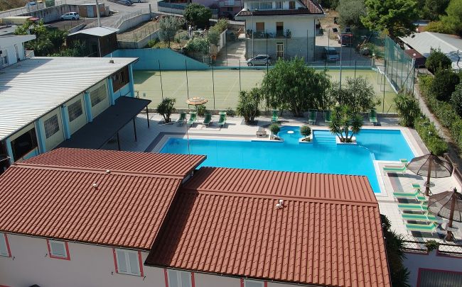 Vista dall'alto di una piscina accanto a edifici con tetto in tegole rosse, vicino a campi sportivi.