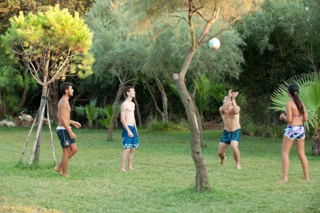 Quattro persone giocano a pallavolo su un prato, circondato da alberi.