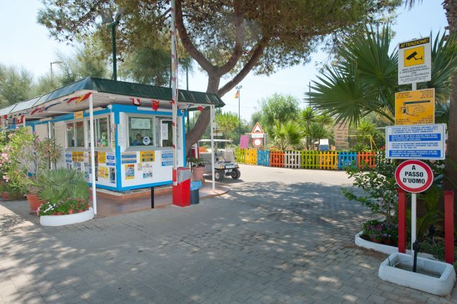 Ingresso con casetta di accoglienza e cartelli, circondato da piante e alberi.