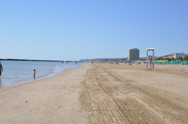 Spiaggia sabbiosa con mare calmo, alcune persone in lontananza e un edificio alto sullo sfondo.