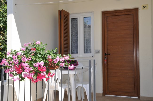 Un piccolo patio con fiori rosa, tavolo bianco con sedie, porta in legno e finestra.