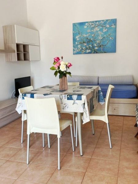 Sala da pranzo con tavolo apparecchiato, sedie bianche e quadro di fiori su sfondo blu.