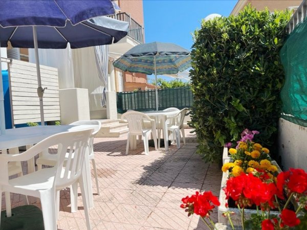 Cortile esterno con sedie bianche, ombrelloni e fiori colorati in vaso.