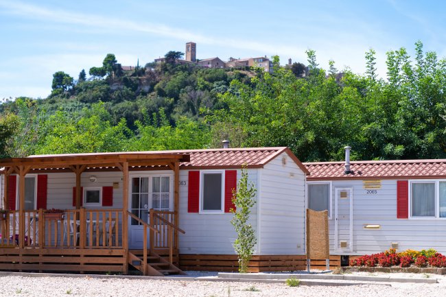 Due case mobili bianche con persiane rosse tra gli alberi, una collina con edifici sullo sfondo.