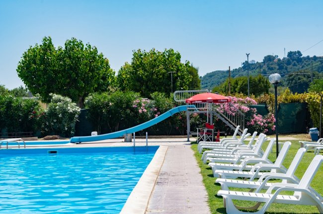 Piscina con scivolo azzurro, sdraio bianche su prato verde e ombrellone rosso.