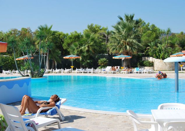 Piscina con donna che prende il sole su un lettino, ombrelloni e palme sullo sfondo.
