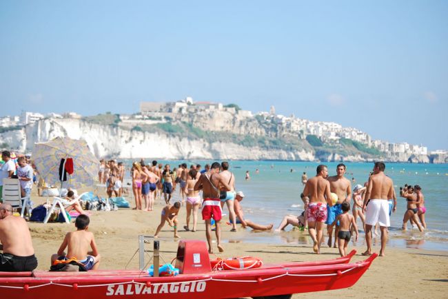 Spiaggia affollata con bagnanti e un pedalò rosso, vista su scogliera e città.