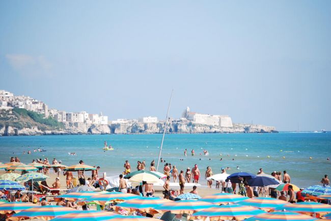 Spiaggia affollata con ombrelloni colorati, mare azzurro e un borgo sullo sfondo.