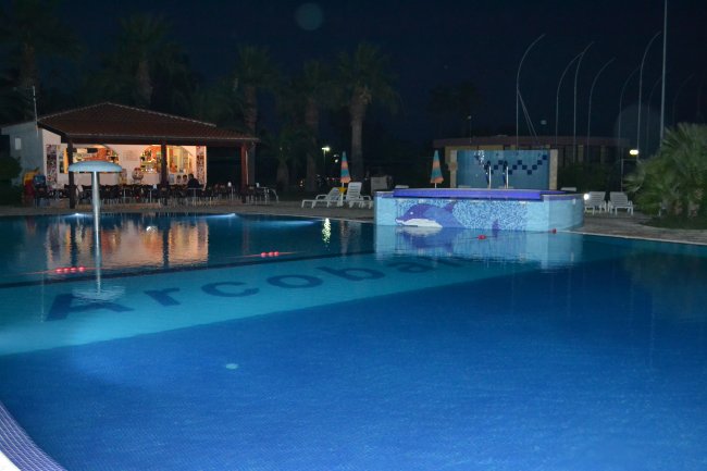 Piscina illuminata di notte con un edificio aperto e palme sullo sfondo.