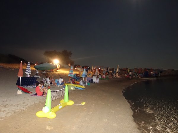 Una spiaggia notturna con persone, ombrelloni colorati e un gioco gonfiabile.