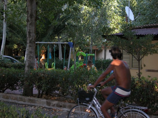 Un uomo in bicicletta passa davanti a un parco giochi con altalene e verde.