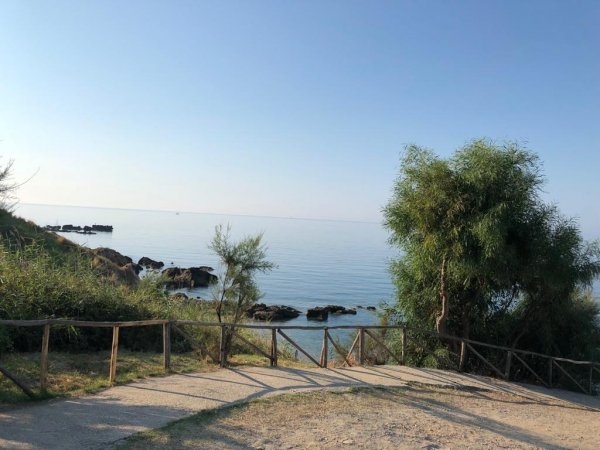 Sentiero sterrato con vista sul mare calmo, circondato da alberi e rocce.