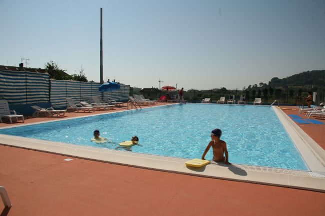 Piscina all'aperto con bambini che nuotano. Lettini e ombrelloni lungo i bordi.