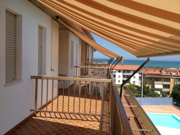 Balcone con tende da sole arancioni e vista su piscina, palazzi e mare.