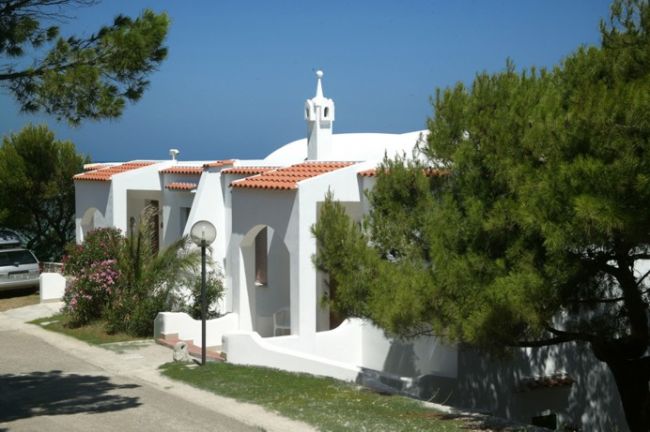 Villa bianca con tetto in tegole rosse, circondata da pini, vista sul mare.