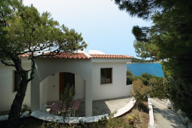 Casa bianca con terrazza coperta, pini intorno e vista sul mare.