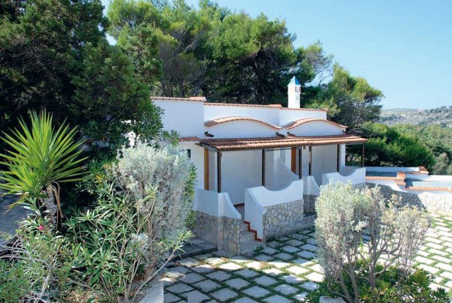 Casa bianca circondata da vegetazione, con pietra e tetto in terracotta, vista esterna.