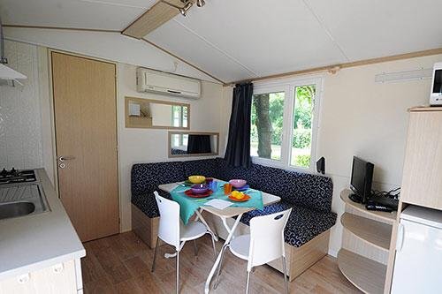 Interno di un bungalow con tavolo da pranzo, cucina e divano; finestra con vista sul verde.