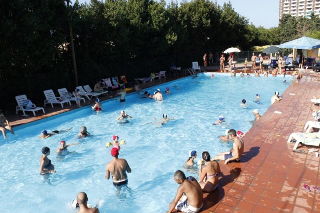 Piscina affollata con persone che nuotano e si rilassano su lettini, circondata da alberi.