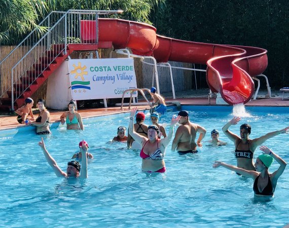 Persone in una piscina vicino a uno scivolo rosso, un cartello delinea il nome del villaggio.