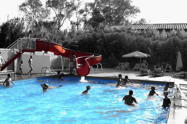 Piscina con scivolo rosso, molte persone nuotano e si divertono sotto il sole.