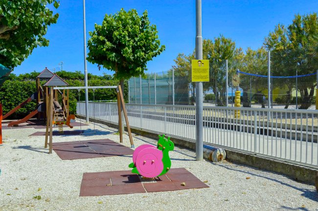 Area giochi con altalene e scivolo, circondata da alberi e recinzioni.