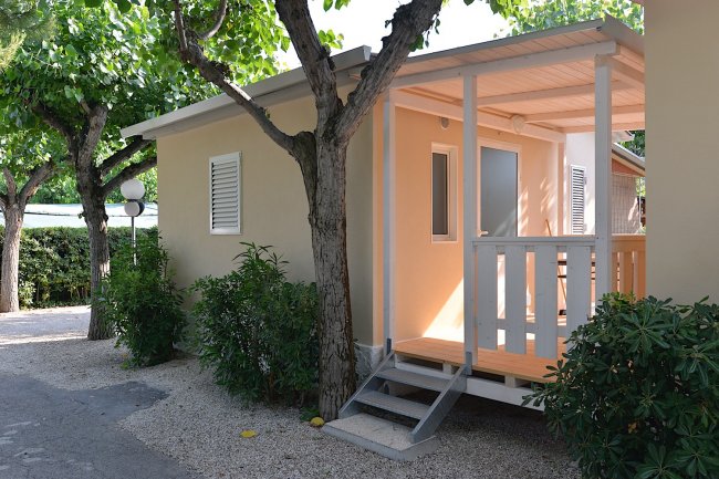 Piccola casa beige con veranda coperta e alberi intorno.