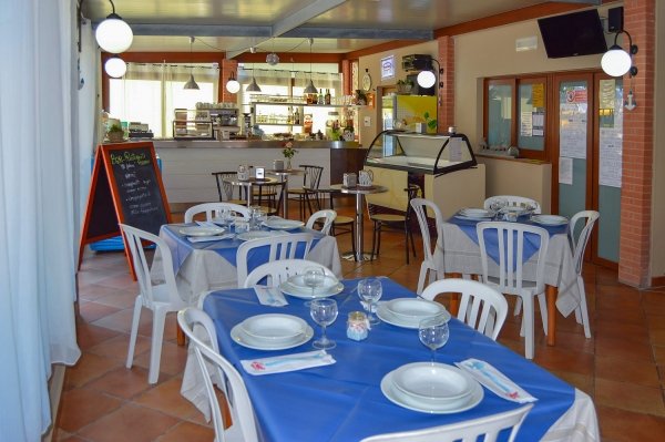 Interno di ristorante con tavoli apparecchiati, sedie in plastica, e vetrina sullo sfondo.
