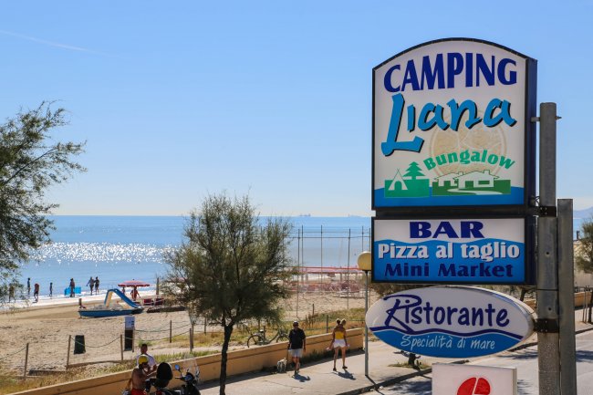 Un cartello di un campeggio vicino a una spiaggia. Persone camminano lungo la strada.