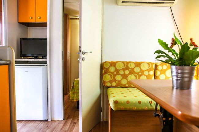 Interno di un bungalow con divano a motivi gialli, tavolo, pianta e cucina con frigorifero.