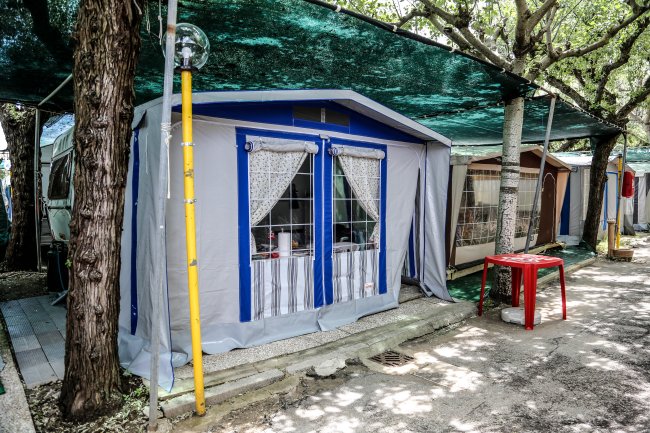 Una tenda da campeggio con finestre blu e tende a pizzo, ombreggiata da alberi.