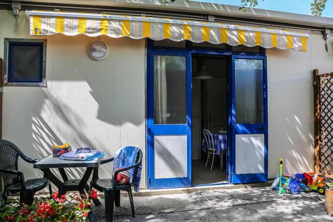 Esterno di una casa con porte blu, tavolo e sedie da giardino, giochi per bambini sul pavimento.