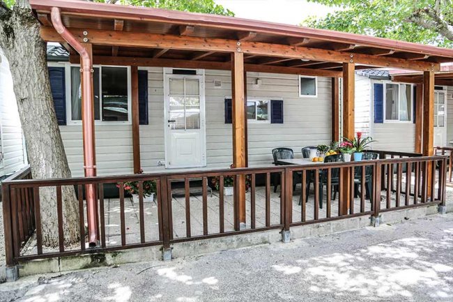 Veranda in legno con tavolo e sedie, circondata da un parapetto e alberi.