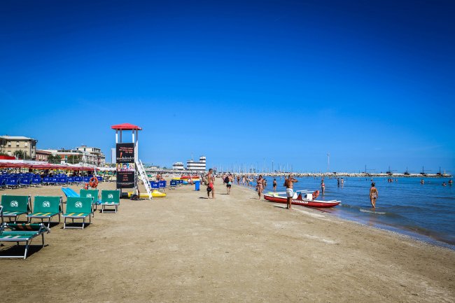 Spiaggia con lettini verdi, torre di salvataggio, alcune persone in acqua e vista sul mare limpido.