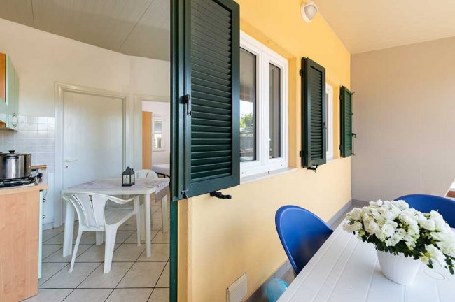 Patio con tavolo e sedie, vista su cucina interna, persiane verdi.