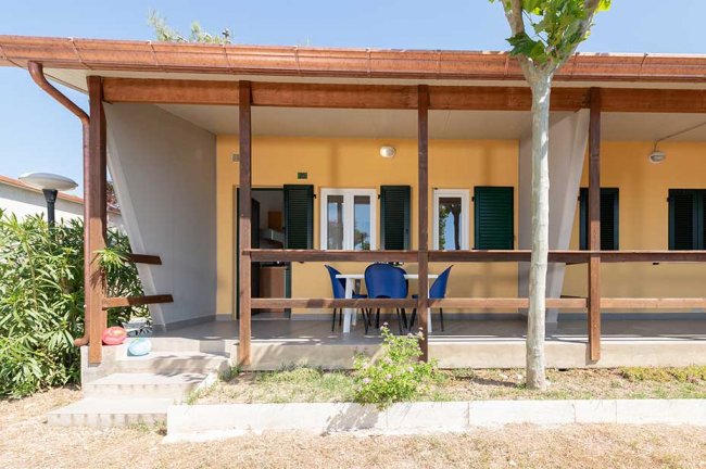 Portico di una casa con tavolo e sedie, circondato da verde.