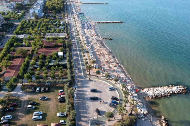 Una strada con spiaggia e mare. Ombrelloni e lettini sulla sabbia, auto parcheggiate a sinistra.