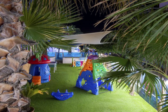 Un parco giochi con scivolo colorato e giochi in plastica su un prato verde circondato da palme.