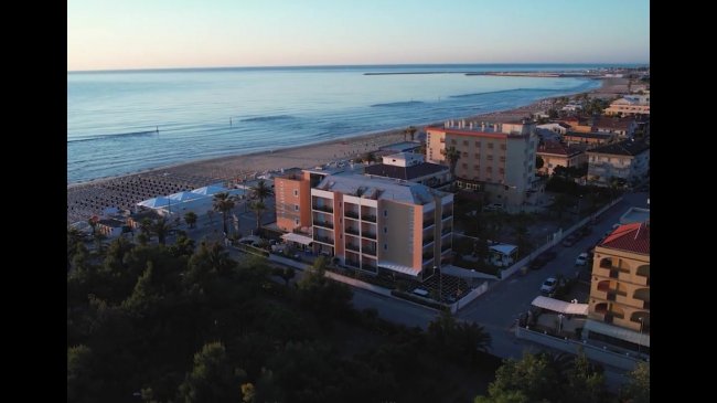 Vista aerea di edifici costieri vicino alla spiaggia al tramonto, con il mare calmo sullo sfondo.