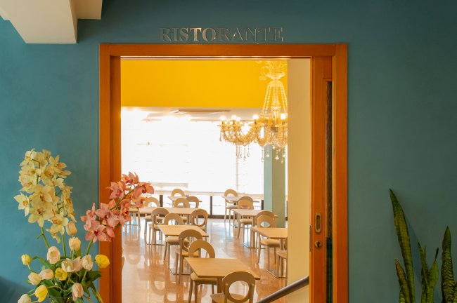 Ingresso di un ristorante con vista su tavoli e sedie, decorazioni floreali e un lampadario.