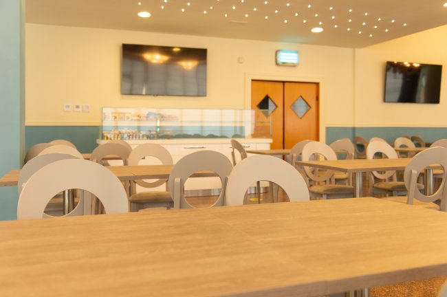 Sala da pranzo con tavoli di legno, sedie dal design moderno e TV a schermo piatto su pareti chiare.