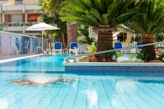 Piscina con palme e lettini blu, in un ambiente soleggiato.