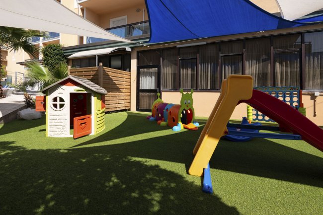 Un'area giochi per bambini con casetta, scivolo colorato e tunnel su erba sintetica.