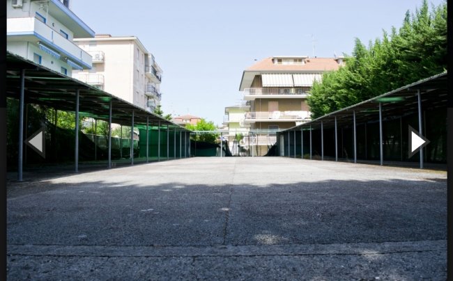 Un parcheggio vuoto con tettoie laterali, circondato da edifici residenziali e alberi.