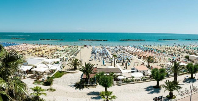 Spiaggia affollata con ombrelloni colorati, palme e mare blu sullo sfondo.
