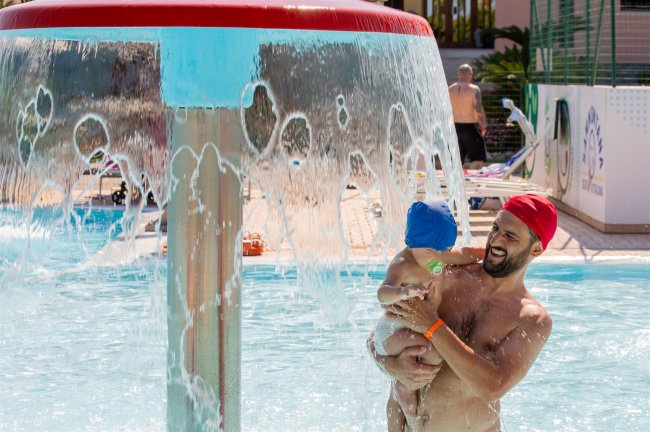 Uomo con bambino sotto una fontana a cascata in una piscina.